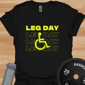 Könnte beinhalten: Ein schwarzes Kurzarm-T-Shirt mit dem gelben Text LEG DAY, der viermal mit einer abgenutzten Textur wiederholt wird. Ein gelbes Rollstuhl-Symbol ist über dem Text zentriert. Das T-Shirt hat einen Rundhalsausschnitt und eine Standardpassform.