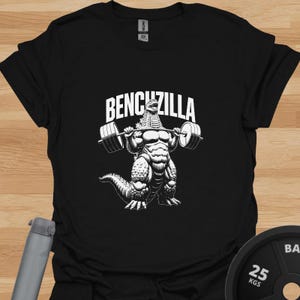 Peut inclure: T-shirt noir avec un motif blanc d'une créature ressemblant à Godzilla soulevant des poids. Le texte "BENCHZILLA" est au-dessus de la créature. Le t-shirt est en matière souple.