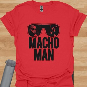 K&ouml;nnte beinhalten: Rotes T-Shirt mit einer schwarzen Grafik von Sonnenbrillen mit zwei Gesichtern und den Worten "MACHO MAN" in Schwarz. Das T-Shirt besteht aus einem weichen Material.
