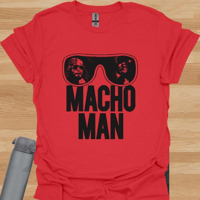 K&ouml;nnte beinhalten: Rotes T-Shirt mit einer schwarzen Grafik von Sonnenbrillen mit zwei Gesichtern und den Worten "MACHO MAN" in Schwarz. Das T-Shirt besteht aus einem weichen Material.