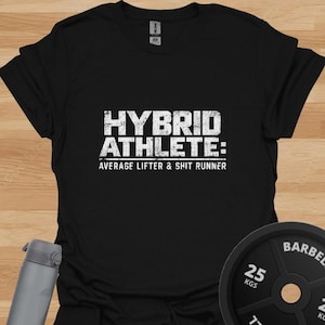 Könnte beinhalten: Ein schwarzes Kurzarm-T-Shirt mit weißem, abgenutztem Text, der HYBRID ATHLETE: AVERAGE LIFTER & SHIT RUNNER lautet. Das T-Shirt hat einen Rundhalsausschnitt und eine Standardpassform, abgebildet mit einer grauen Trinkflasche und einer 25 kg Hantelscheibe.