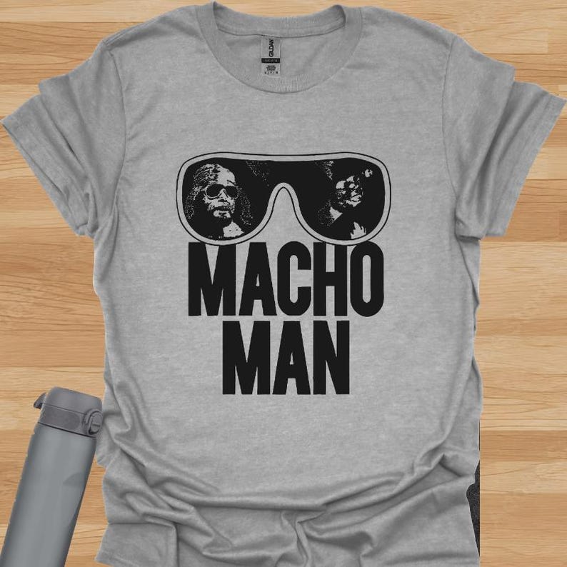 K&ouml;nnte beinhalten: Ein hellgraues T-Shirt mit einer schwarzen Grafik von Sonnenbrillen mit zwei Gesichtern und den Worten "MACHO MAN" in fetten, schwarzen Buchstaben. Das Shirt ist aus einem weichen Material.