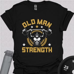 Black unisex gym t shirt featuring a bold gold and white “Old Man Strength” design with a muscular lifter holding a barbell overhead, surrounded by stars and laurel wreaths, laid flat with gym accessories in the background.