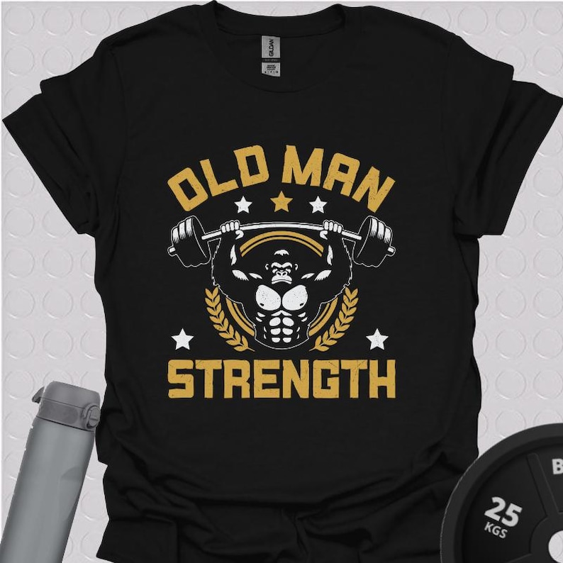 Black unisex gym t shirt featuring a bold gold and white “Old Man Strength” design with a muscular lifter holding a barbell overhead, surrounded by stars and laurel wreaths, laid flat with gym accessories in the background.