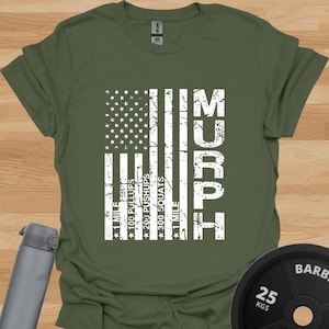 Könnte beinhalten: Olivgrünes T-Shirt mit einem weißen, verblassten Design der amerikanischen Flagge und dem Wort "MURPH". Die Grafik enthält Trainingsdetails: 1 Meile Laufen, 100 Klimmzüge, 200 Liegestütze, 300 Kniebeugen und 1 Meile Laufen.