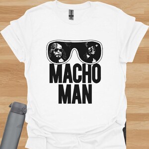 K&ouml;nnte beinhalten: Wei&szlig;es T-Shirt mit einer schwarz-wei&szlig;en Grafik einer Sonnenbrille mit zwei Gesichtern und den Worten "MACHO MAN" in fetten, schwarzen Buchstaben. Das Shirt ist aus einem weichen Material.