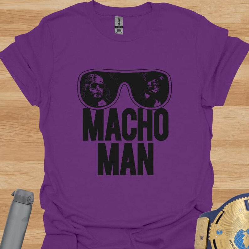 K&ouml;nnte beinhalten: Ein lila T-Shirt mit einer schwarzen Grafik. Die Grafik zeigt eine Sonnenbrille mit zwei Gesichtern im Inneren und die Worte "MACHO MAN" in Schwarz. Das T-Shirt ist einfarbig und scheint aus Baumwolle zu sein.