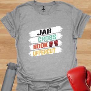 May include: Grey t-shirt with a white graphic that says "JAB CROSS HOOK UPPERCUT" with red boxing gloves on the "HOOK" line.