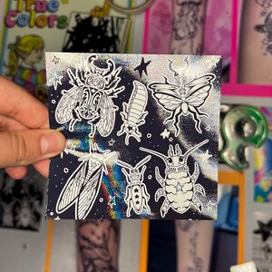 May include: A small, square sticker sheet featuring black and white illustrations of various insects, including beetles and butterflies, against a dark blue background with a holographic, glittery effect. Stars are scattered around the insects.