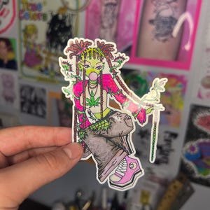 Peut inclure: Un autocollant coloré représentant un personnage de dessin animé à la peau verte, aux cheveux roses et une bulle de chewing-gum. Le personnage porte un crop top avec une feuille de cannabis, un pantalon marron et des baskets roses. L'autocollant a une bordure blanche.