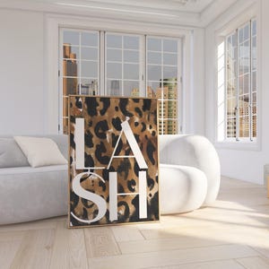 May include: A framed artwork featuring a leopard print background with the word "LASH" in large white capital letters. The artwork is displayed in a bright, modern room with a light-colored sofa and chair.