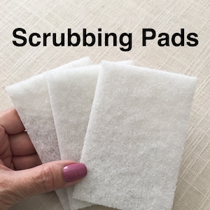 May include: Three white scrubbing pads, made of a soft, textured material. The pads are rectangular and are held in a hand.