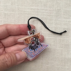 May include: A clear acrylic charm featuring a vintage sewing machine on a patterned rug. The charm has a black string attached for hanging.