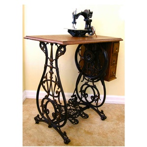 Peut inclure: Table de machine &agrave; coudre antique en fonte noire avec un plateau en bois et un ensemble de tiroirs sur le c&ocirc;t&eacute;. La table a des volutes orn&eacute;es et une grande roue sur le c&ocirc;t&eacute;.