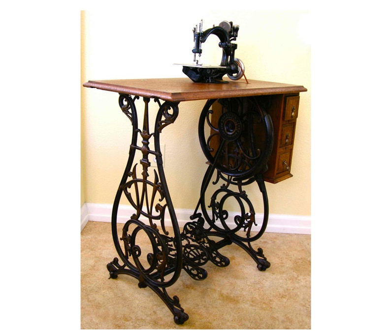 Peut inclure: Table de machine &agrave; coudre antique en fonte noire avec un plateau en bois et un ensemble de tiroirs sur le c&ocirc;t&eacute;. La table a des volutes orn&eacute;es et une grande roue sur le c&ocirc;t&eacute;.