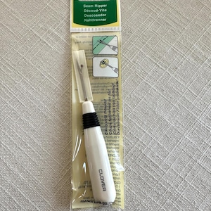 May include: A white seam ripper with a black handle and a silver blade. The packaging is green and white and says "Clover Seam Ripper" in English, French, Spanish, and German.