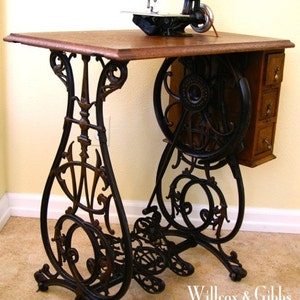 Puede incluir: M&aacute;quina de coser antigua de pedal negro con una mesa de madera y patas de metal ornamentadas. La m&aacute;quina tiene un mecanismo de puntada de cadena y est&aacute; etiquetada como "Willcox & Gibbs 1903 Chain stitch sewing machine".
