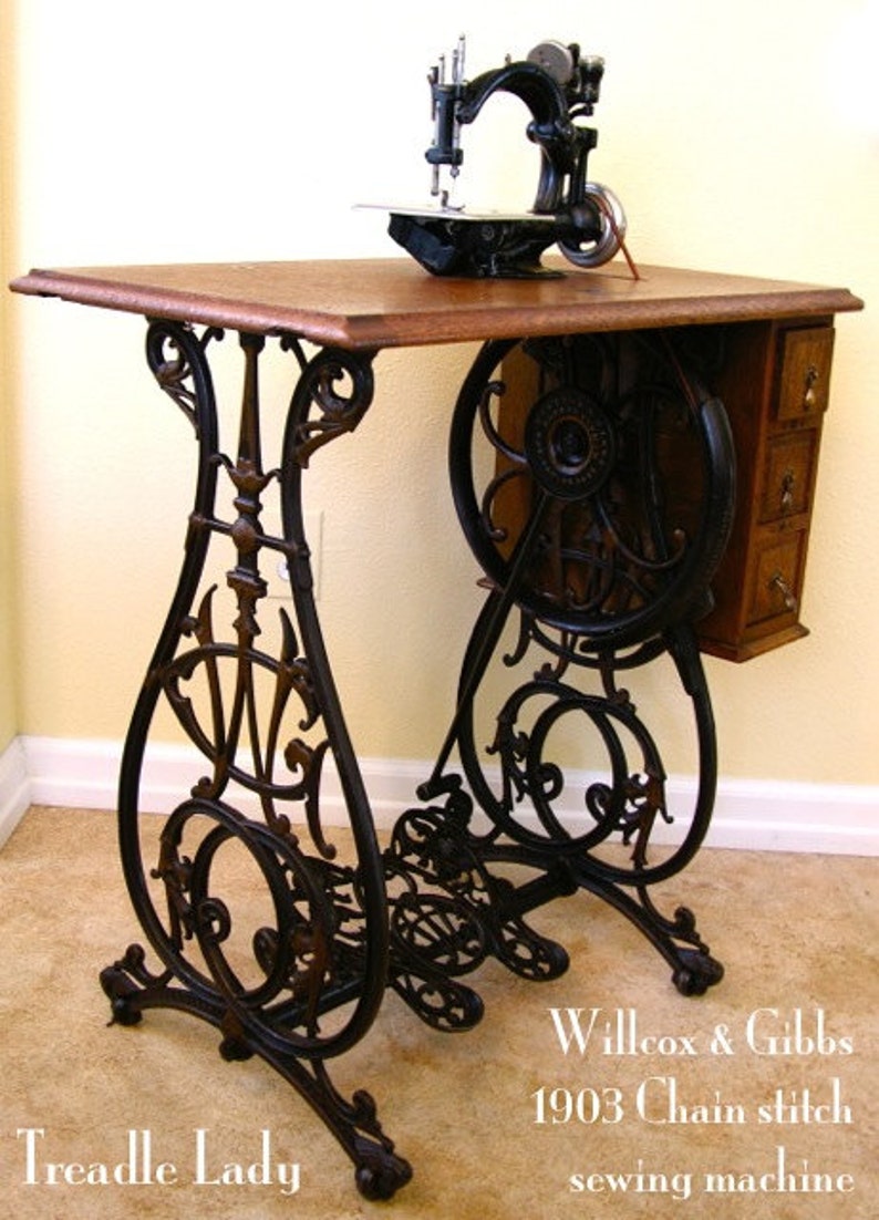 Puede incluir: M&aacute;quina de coser antigua de pedal negro con una mesa de madera y patas de metal ornamentadas. La m&aacute;quina tiene un mecanismo de puntada de cadena y est&aacute; etiquetada como "Willcox & Gibbs 1903 Chain stitch sewing machine".