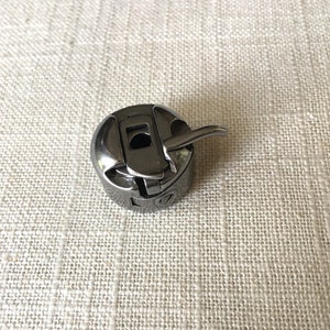 May include: A black metal bobbin case for a sewing machine. The bobbin case has a small lever on the side.
