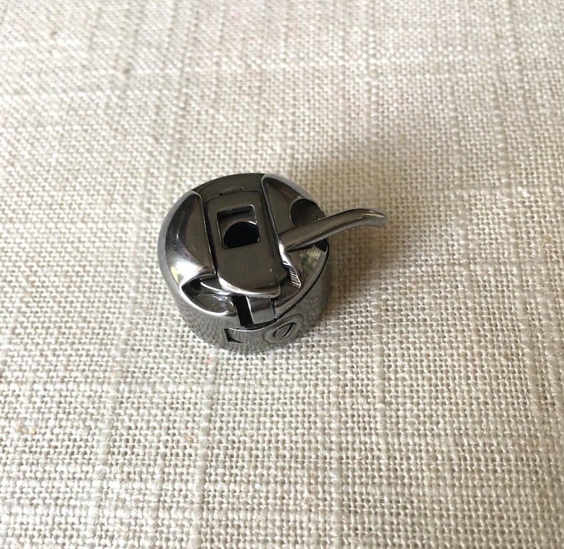 May include: A black metal bobbin case for a sewing machine. The bobbin case has a small lever on the side.