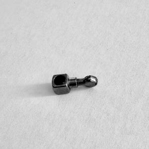 May include: A small, metallic, dark gray object with a square base and a rounded end. The object has a cylindrical connecting piece between the square base and the rounded end. The square base has a hole in the center.