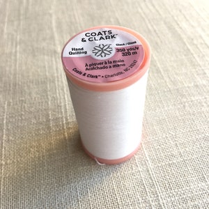 May include: A spool of white Coats & Clark hand quilting thread. The spool has a pink and white label with the text "COATS & CLARK" and "Hand Quilting". The label also indicates 350 yards/v or 320 meters of thread.