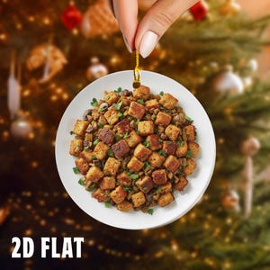 May include: A white, round Christmas ornament featuring a plate of stuffing with croutons, mushrooms, and parsley. The ornament is held by a gold string against a blurred Christmas tree background. The text "2D FLAT" is at the bottom.