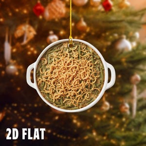 May include: A white, round Christmas ornament shaped like a casserole dish filled with green beans and crispy fried onions. The ornament is hanging in front of a blurred Christmas tree. The text "2D FLAT" is at the bottom.