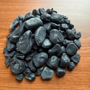 May include: A pile of smooth, dark gray stones of varying sizes. The stones have a natural, weathered appearance and are arranged on a wooden surface. The stones are a solid, neutral color.