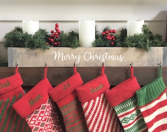 May include: A Christmas mantel display with a wooden box holding candles, greenery, and red berries. The box reads "Merry Christmas." Red stockings hang below, each with a name. Decorative items include a green box, a gift, and wooden trees.