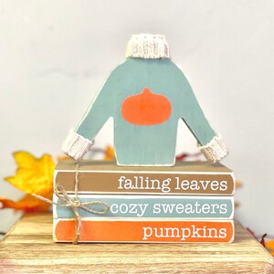 May include: A stack of three wooden blocks with the words "falling leaves", "cozy sweaters", and "pumpkins" painted on them. A wooden cutout of a blue sweater with a pumpkin on the front is on top of the blocks.