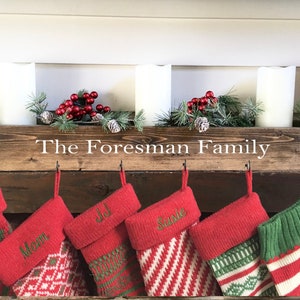 May include: A wooden box with the text "The Foreman Family" on the front, decorated with greenery and red berries. Five stockings are hung on hooks below the box, each with a name embroidered on the cuff: Dad, Mom, Jill, Susie, and a green stocking with white stripes.