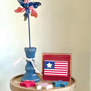 May include: A patriotic display featuring a wooden pinwheel with red and white stripes and blue stars, a blue candle holder, a red block with an American flag, and star-shaped decorations. The items are arranged on a wooden pedestal.