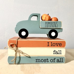May include: A blue wooden truck carrying three orange pumpkins in its bed. The truck is sitting on top of three stacked books. The books are painted in shades of orange, white, and blue. The top book has the text "I love fall most of all" written on it.