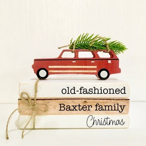 May include: A stack of three white books with red and black lettering. The top book says "old-fashioned", the middle book says "Baxter family", and the bottom book says "Christmas". A red toy car with a small green Christmas tree on top sits on the books.