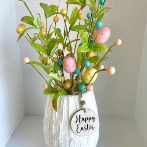 May include: A white knit vase holds an Easter arrangement. The arrangement includes green leaves, colorful speckled eggs, and small round ornaments in yellow, pink, and blue. A wooden tag with "Happy Easter" hangs from the vase.