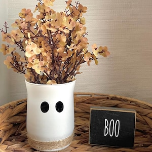 May include: A white vase with black eyes and a jute-wrapped base, filled with peach-colored flowers. A small black wooden sign with the word "BOO" in white sits nearby. The arrangement is in a woven basket, perfect for Halloween decor.