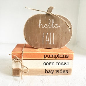 May include: A wooden pumpkin sign with the text "hello fall" in white lettering, sitting on top of a stack of three books. The books are painted in shades of orange, white, and brown, and each has a different fall-themed word written on it: "pumpkins", "corn maze", and "hay rides".
