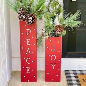 May include: Two red wooden planters with white polka dots and the words "PEACE" and "JOY" painted on them. The planters are filled with green pine branches and pine cones.