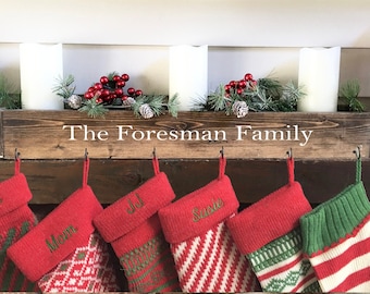 May include: A wooden box with the text "The Foreman Family" on the front, decorated with greenery and red berries. Five stockings are hung on hooks below the box, each with a name embroidered on the cuff: Dad, Mom, Jill, Susie, and a green stocking with white stripes.