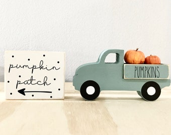 May include: A small wooden truck painted light blue with white wheels, carrying two orange pumpkins. The truck has a sign on the back that says "PUMPKINS". A white wooden block with black polka dots and the text "pumpkin patch" with a black arrow pointing left is in the background.