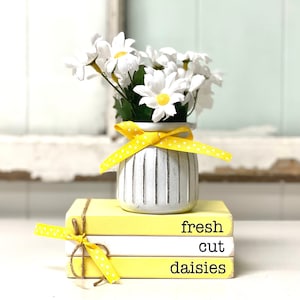 May include: A stack of three yellow books with white accents. The top book says "fresh cut daisies" in black text. A white vase with yellow polka dot ribbon holds a bouquet of white daisies.