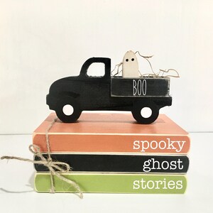 May include: A black wooden truck with a white ghost in the bed. The truck is sitting on top of three stacked books. The books are orange, black, and green. The books have the words "spooky ghost stories" written on them.