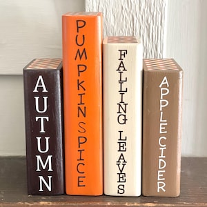 May include: Four wooden blocks painted in autumn colors, arranged to resemble books. The blocks are labeled "AUTUMN", "PUMPKIN SPICE", "FALLING LEAVES", and "APPLE CIDER" in white lettering. The tops of the blocks have a brown and white pattern.