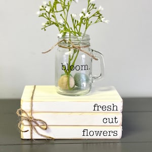 May include: A stack of four white books with the words "fresh cut flowers" written on the bottom book. A mason jar with the word "bloom." written on it is sitting on top of the books. The jar is filled with white flowers.