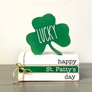 May include: A stack of three white books with green accents. The top book says "happy", the middle book says "St. Patty's", and the bottom book says "day". A green shamrock cutout with the word "LUCKY" written on it sits on top of the books.