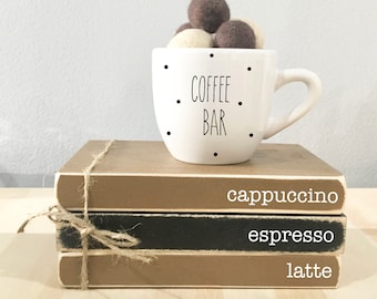 May include: A white ceramic coffee mug with black dots and the text "COFFEE BAR" on it. The mug is filled with brown and white felt balls and is sitting on top of a stack of three books. The books are labeled "cappuccino", "espresso", and "latte".