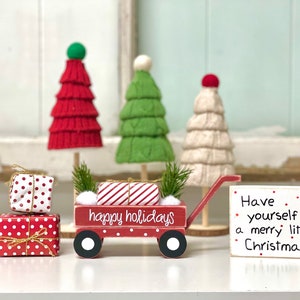 May include: Red wooden wagon filled with wrapped gifts and a sign that reads "Happy Holidays".  The wagon is decorated with a green sprig and white pom poms.  There is a sign next to the wagon that reads "Have yourself a merry little Christmas".