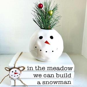 May include: A white snowman-shaped vase with black eyes, a red carrot nose, and a green plant with red and yellow ornaments. The vase sits atop white books with the text "in the meadow we can build a snowman."