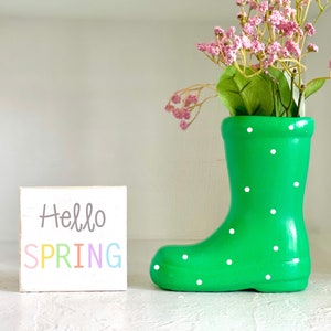 May include: A green rain boot-shaped vase with white polka dots, filled with pink flowers and greenery. A small white wooden block with the words "Hello SPRING" in pastel colors sits beside the vase. The items are displayed on a white surface.
